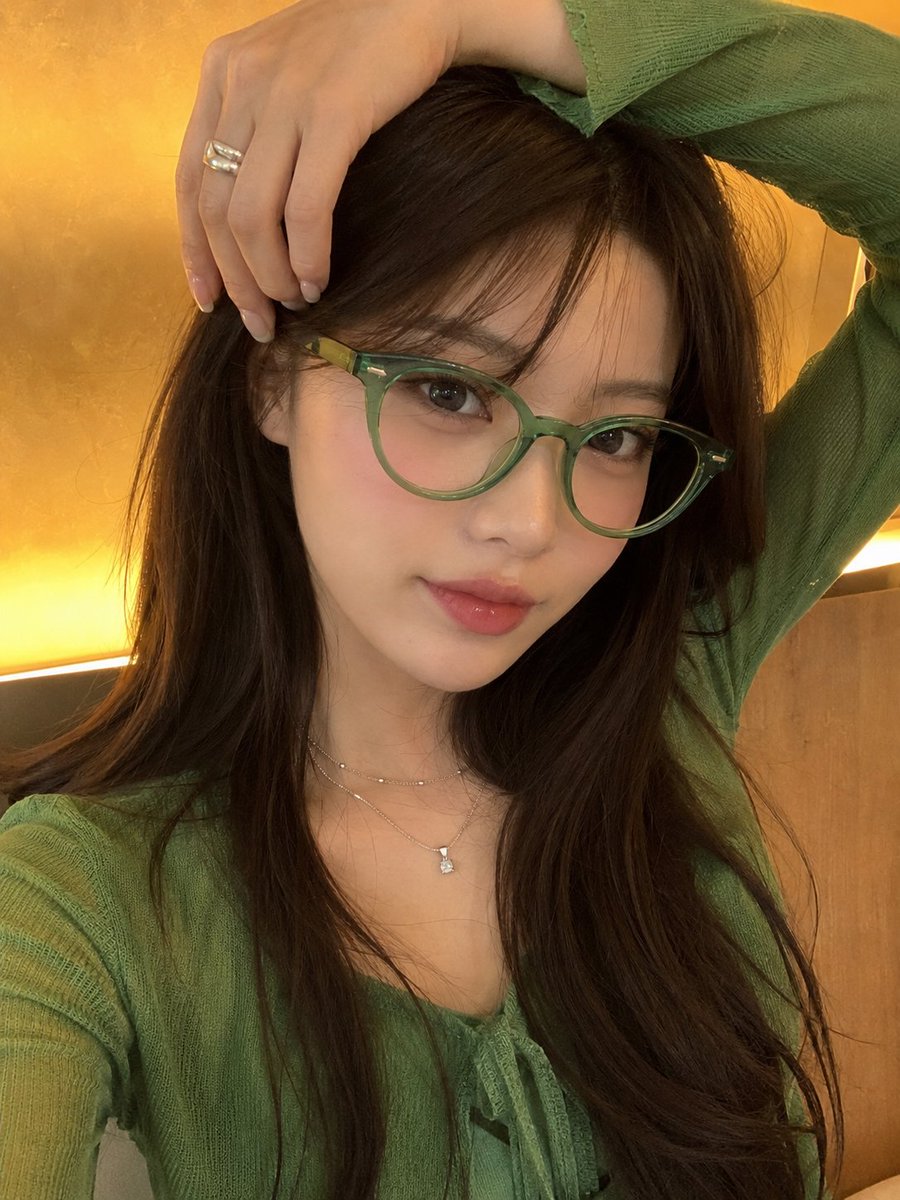 一张高品质的竖版人像照，主体是一位年轻女性，留着长直深棕色头发，戴着时尚的半透明翡翠绿猫眼眼镜，身穿鲜艳的绿色服装。她在室内摆姿势，一只手自然举...
