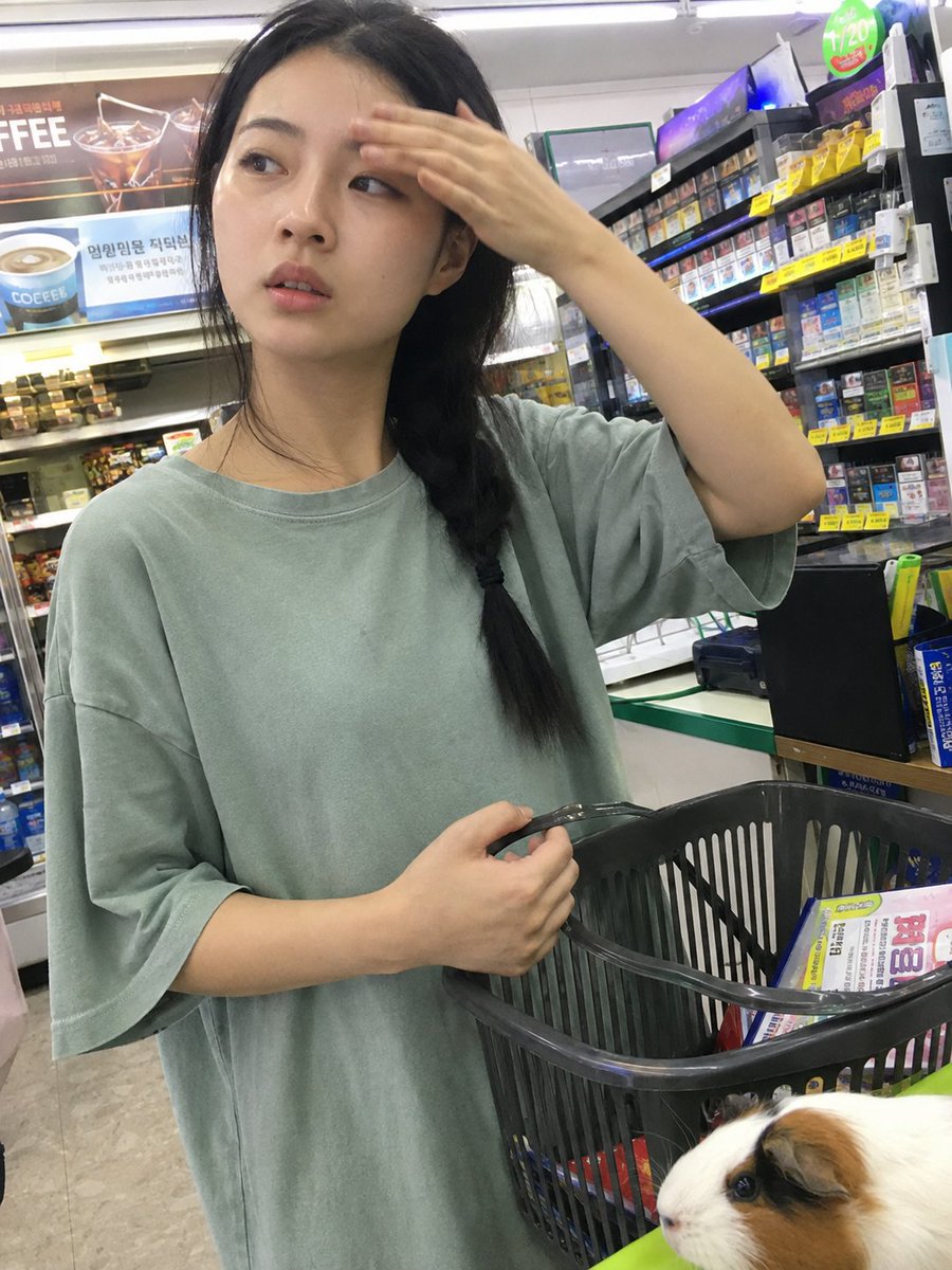 用iPhone前置摄像头意外抓拍的快照，拍摄于一家小型社区便利店内部。一位年轻的韩国女性站在收银台前排队，一边随意地用手抹去额头上的一丝汗珠，一边挎着购物篮。她目光望向一侧，表情略显不耐烦与心...