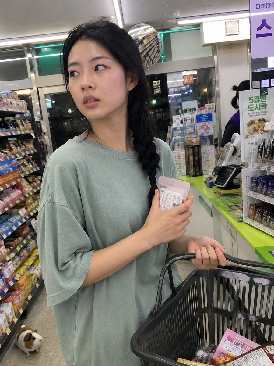 用iPhone前置摄像头意外抓拍的快照，拍摄于一家小型社区便利店内部。一位年轻的韩国女性站在收银台前排队，一边随意地用手抹去额头上的一丝汗珠，一边挎着购物篮。她目光望向一侧，表情略显不耐烦与心...