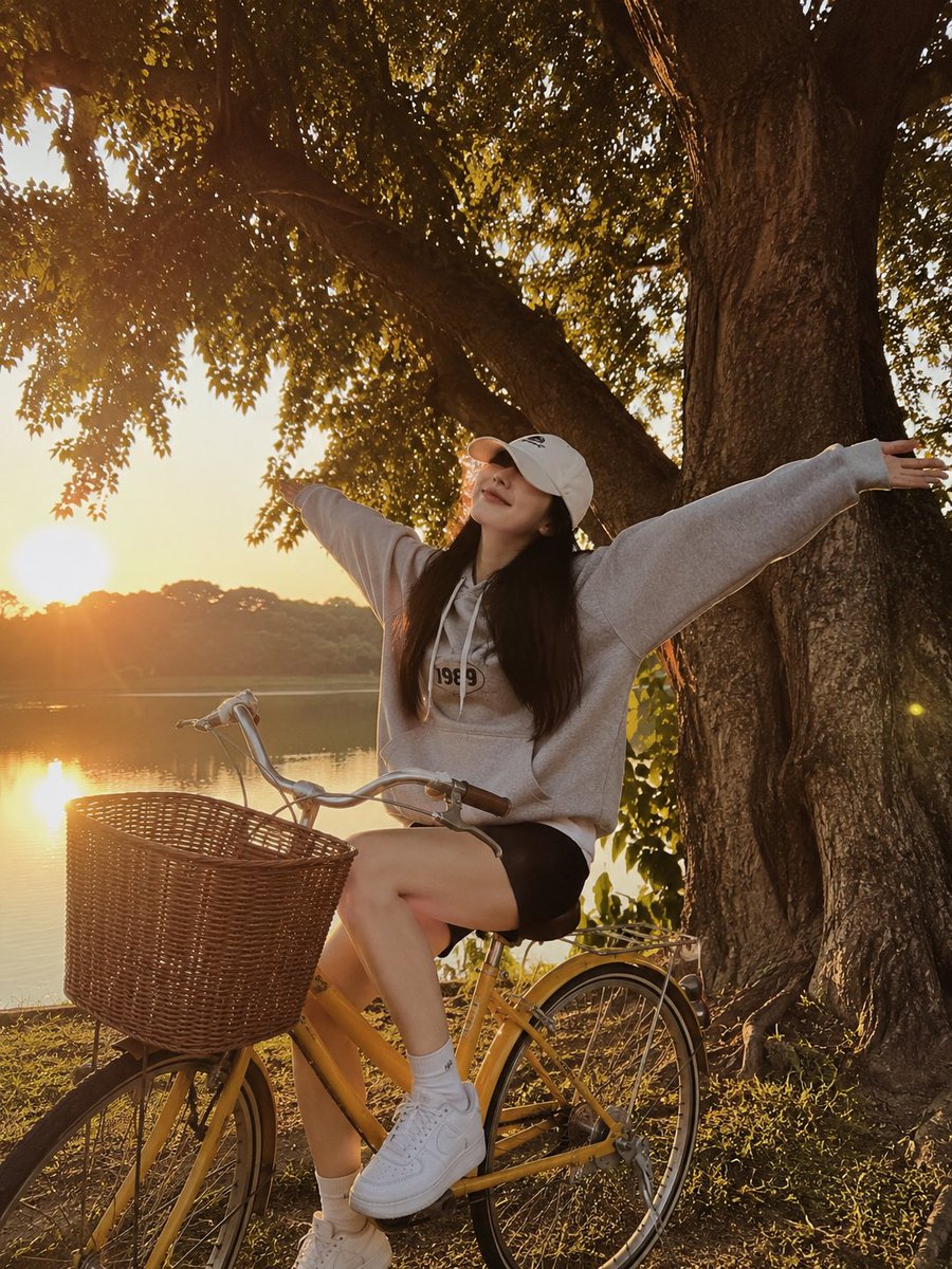 一张黄金时段的写实生活方式摄影照片，画面中一位年轻女子俏皮地坐在一辆复古黄色自行车旁，旁边是一片宁静的湖泊。她张开双臂，摆出愉悦、无忧无虑的姿态，嘴角带着柔和的微笑，脸部被一顶白色棒球帽部分遮...
