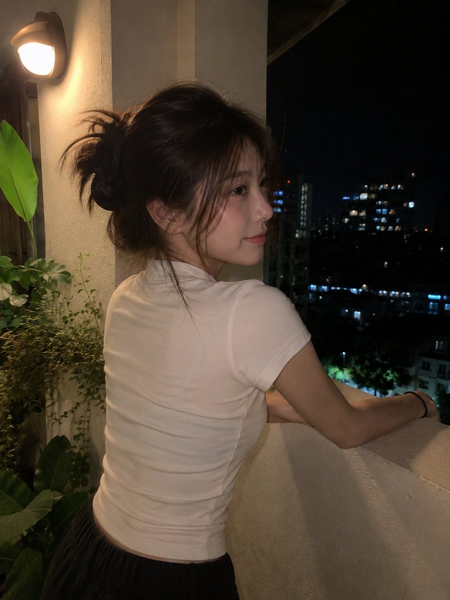 使用 GPT image 2 创建 提示词 一张看起来真实的夜间手机照片：一位年轻的东亚女性随意地站在小型公寓阳台或露台上,由朋友用现代智能手机随手抓拍。她留着中长的深棕色头发,松松地扎成一个...