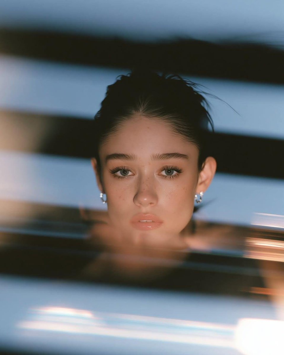 超写实电影感特写肖像：一位美丽的年轻女子，光滑的深色头发梳成整洁的低发髻，浓密的自然眉毛，淡淡的雀斑，裸色亮泽唇彩，带有逼真毛孔的无瑕肌肤，佩戴小巧的银色圈形耳环，身着极简黑色服装。她神情平静...