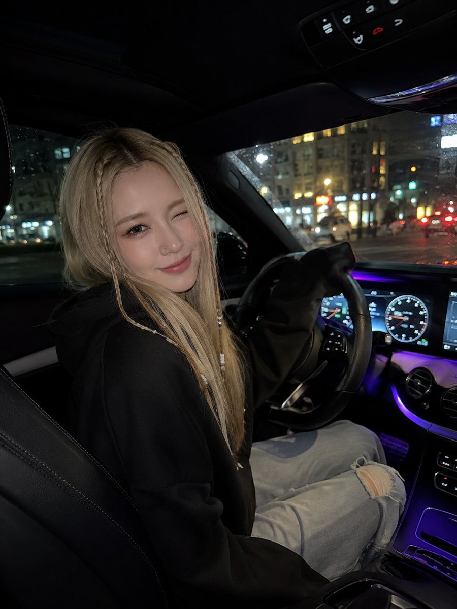 一张超写实的夜间车内自拍，年轻女子坐在驾驶座上，微微转向镜头，调皮地眨眼并带着温柔的微笑。她有一头长直的金发，脸颊两侧各编着精致的细辫，辫子上装...