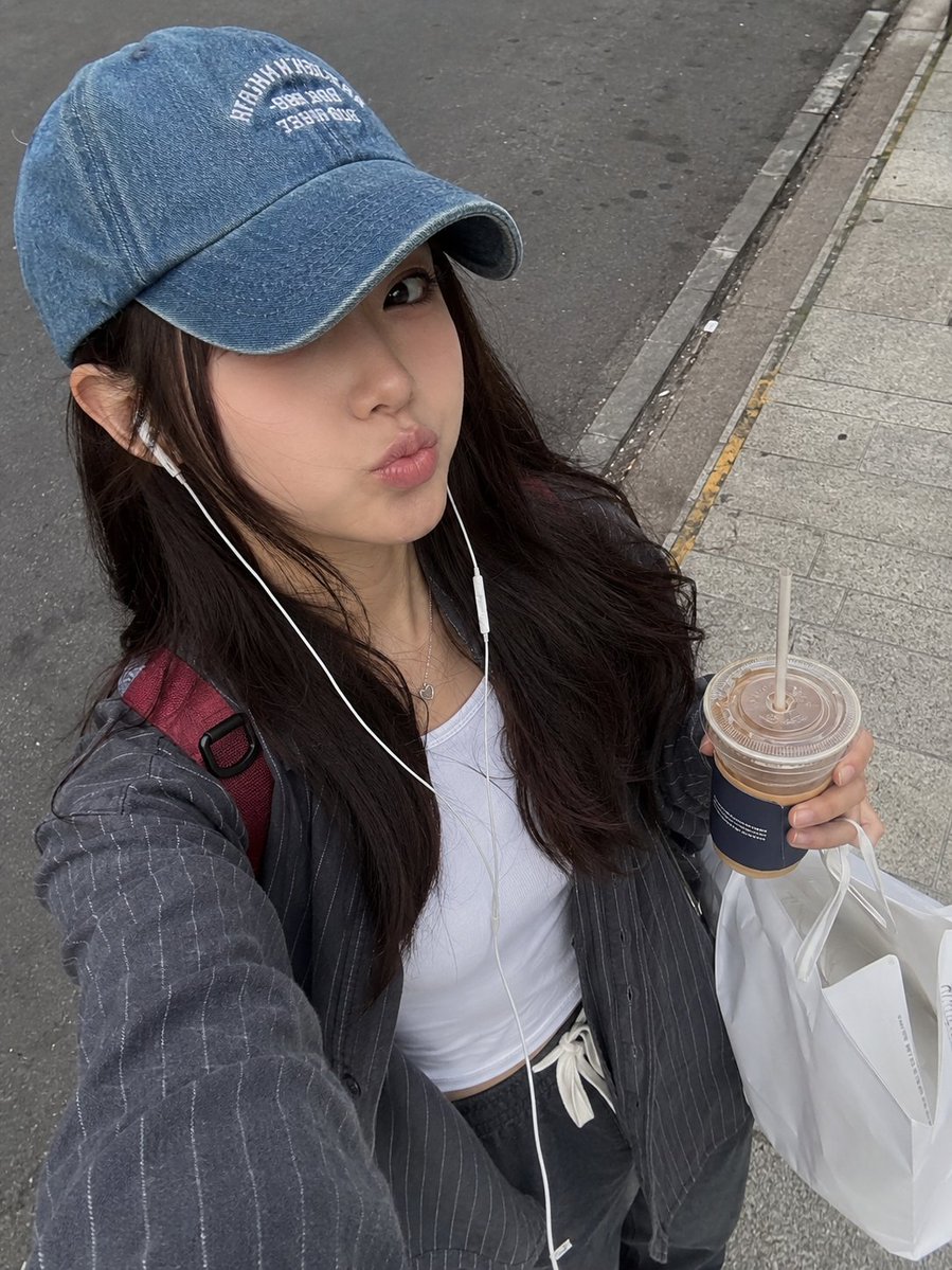 一张照片般逼真的街头风格抓拍自拍照，画面中一位年轻女性正在城市人行道上户外行走。她有一头深棕色长发，自然地垂落在褪色蓝色牛仔棒球帽下，帽子带有白...
