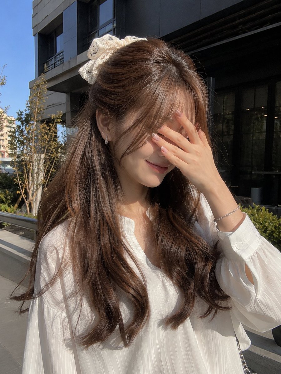 一张年轻女性的户外写实主义肖像照，她站在现代建筑外的明亮自然阳光下。她有一头浓密蓬松的浅棕色长发，呈现出柔和流畅的波浪卷，发帘式刘海轻盈地修饰着...