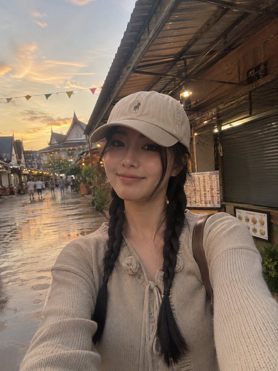 一张照片级写实的竖版旅行自拍照：一位年轻女性在黄金时段身处热闹的集市或旅游村落户外。她戴着米色棒球帽，帽子带有低调的刺绣标志；乌黑长发编成两条整...