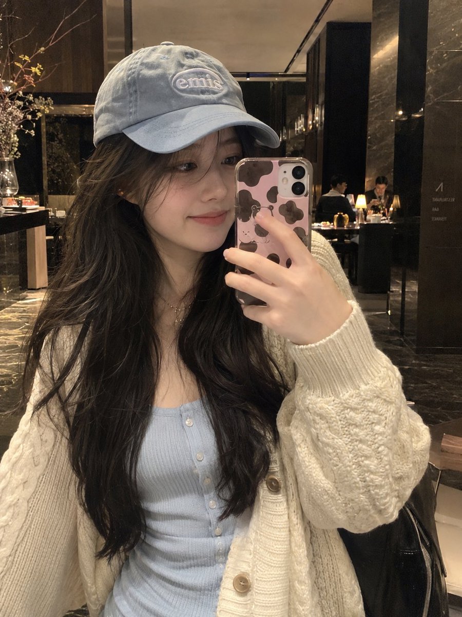 超写实自然智能手机自拍，竖向 3:4，一位年轻东亚女性的室内镜面式随性自拍，背景为一家温馨高档的餐厅或酒店大堂酒廊。 她有着长而自然波浪卷的深棕...