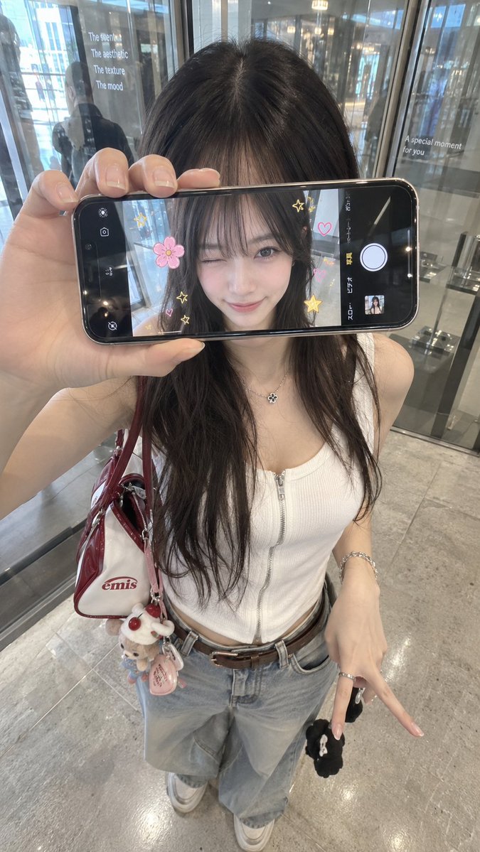 超写实 iPhone 创意人像，身份特征与参考图像完全一致。年轻女性站在室内玻璃店面或反光墙前，从略高的正面角度拍摄。她将一部智能手机水平举在正...