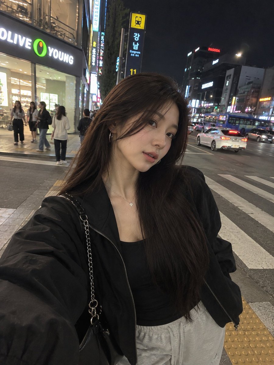 一张照片级写实风格的夜间街头人像照,一位年轻女子在繁华的韩国城市街区中随意地自拍。她留着长而顺直的黑发,妆容柔和自然,佩戴着低调的环形耳环和一条精致的项链。她身穿黑色修身内搭,外搭一件宽松的黑...