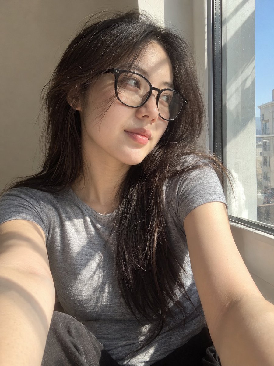 超写实风格的休闲智能手机自拍照：一位年轻东亚女性坐在安静房间内靠近阳光照射的窗户旁。保持与面部参考的高度相似性，包括柔和轮廓的鹅蛋脸、温和的下颌线、自然比例的鼻子、真实的唇形、均衡的眼距、微妙...