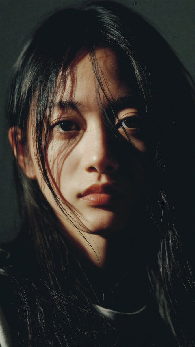 特写电影感人像：一位年轻的东亚女性，长长的略带凌乱的黑发披在肩上，几缕散发拂过脸庞。她直视镜头，表情平静、专注而略带忧郁。左侧柔和而戏剧性的侧光照亮她半边脸庞，另一半则陷入深重的阴影。背景近乎...