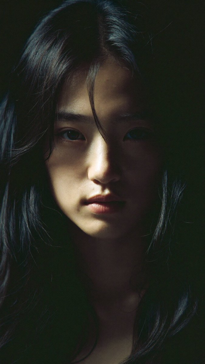 特写电影感人像：一位年轻的东亚女性，长长的略带凌乱的黑发披在肩上，几缕散发拂过脸庞。她直视镜头，表情平静、专注而略带忧郁。左侧柔和而戏剧性的侧光照亮她半边脸庞，另一半则陷入深重的阴影。背景近乎...