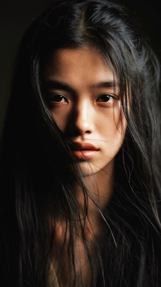 特写电影感人像：一位年轻的东亚女性，长长的略带凌乱的黑发披在肩上，几缕散发拂过脸庞。她直视镜头，表情平静、专注而略带忧郁。左侧柔和而戏剧性的侧光照亮她半边脸庞，另一半则陷入深重的阴影。背景近乎...
