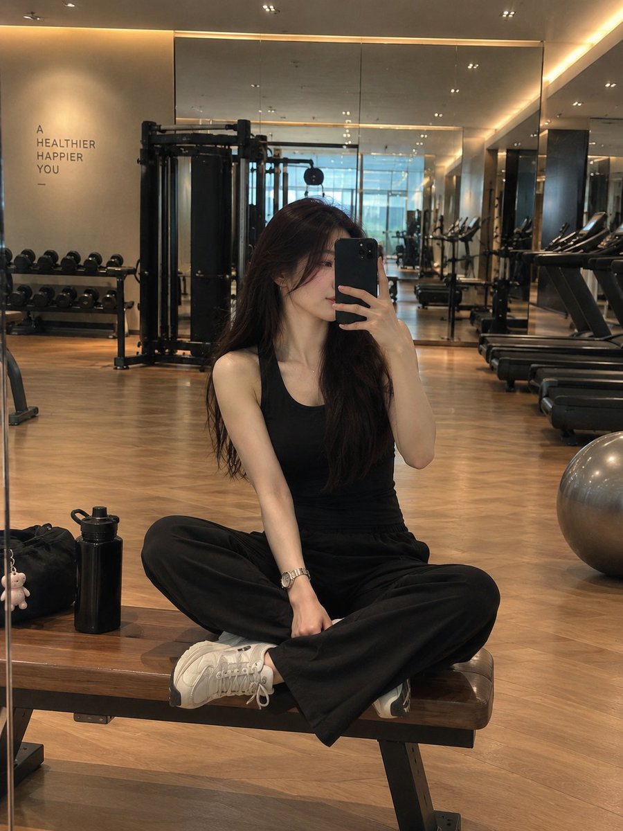 一位时尚年轻女性在现代豪华健身房内对着镜子自拍，盘腿坐在一张木质健身凳上。她留着长而顺直的深棕色头发，神情平静自然，手持一部黑色智能手机置于面前。她身穿一套精致的全黑色无袖运动装，搭配宽松的黑...
