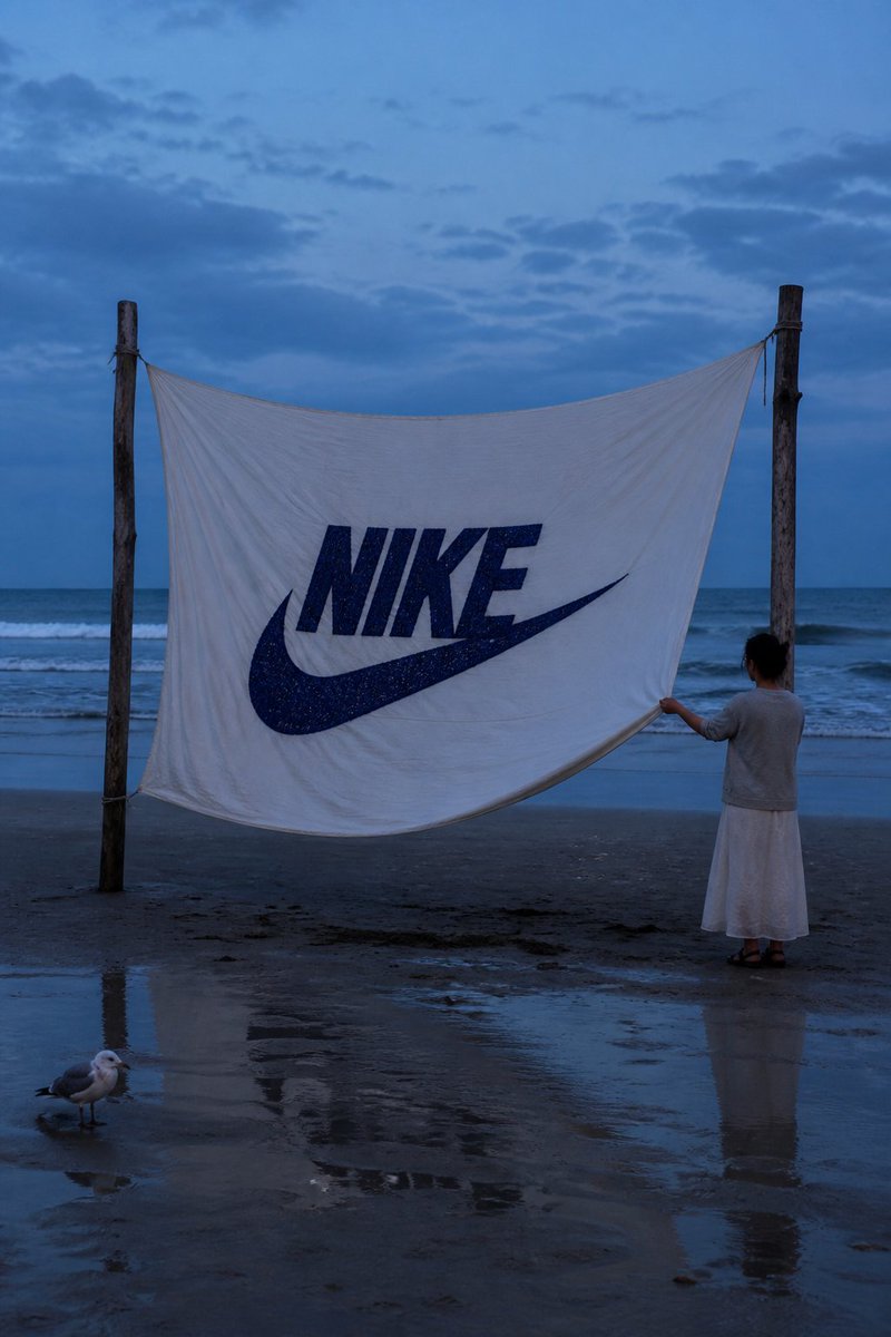 一张写实风格的海岸摄影作品。在蓝调时刻的海滩上，两根浮木杆之间悬挂着一块巨大的白布，中央用粗实的深蓝色线绣有 "NIKE" 字样及其标志，布料随...